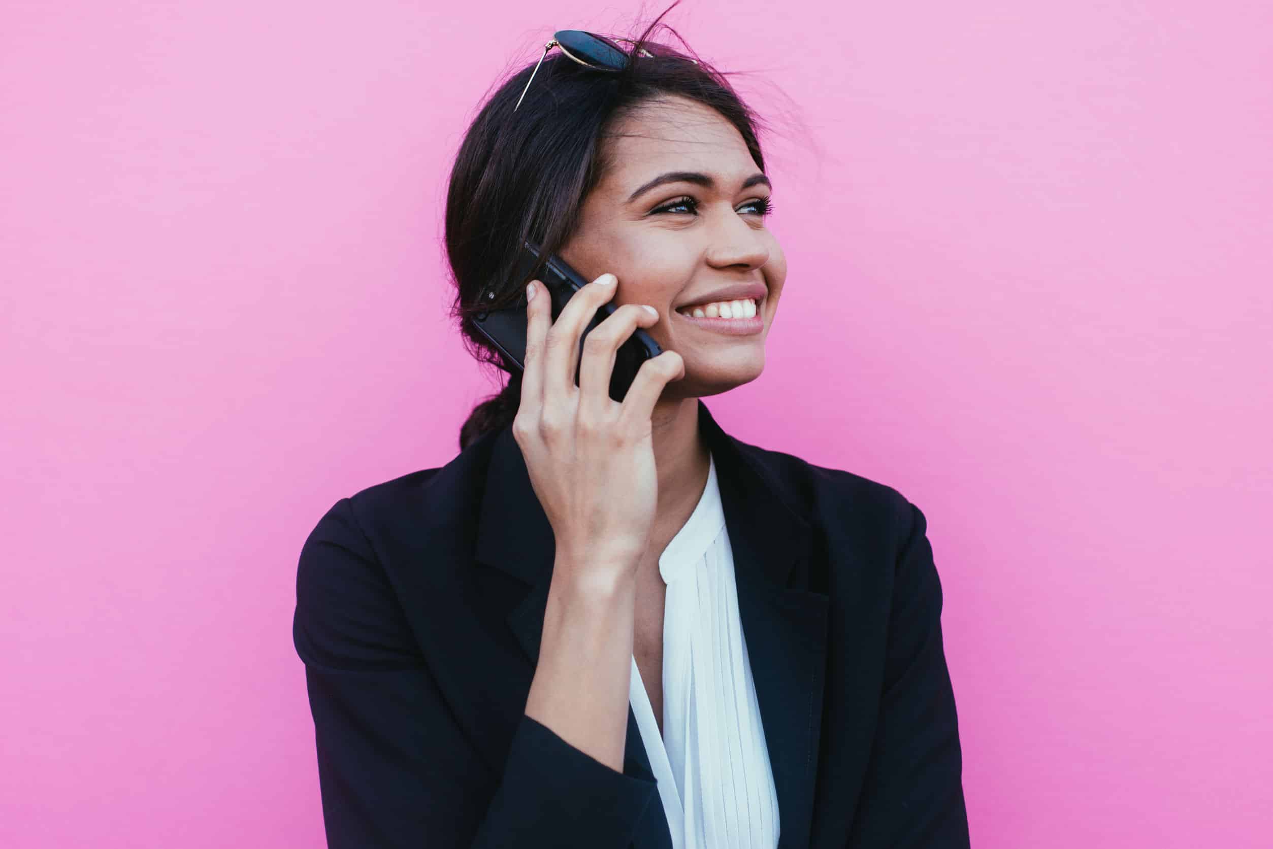 businesswoman smiling while on the phone, standing in front of a pink background