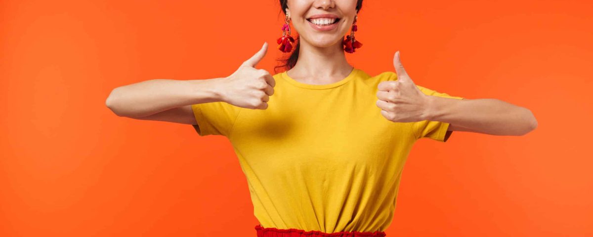 a happy young woman with two thumbs up on against an orange wall background