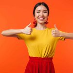 a happy young woman with two thumbs up on against an orange wall background