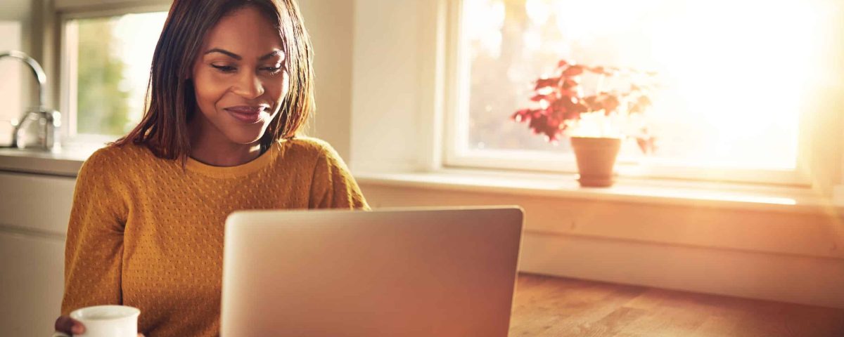 Woman smiling while drinking coffee and sitting with her laptop in the kitchen