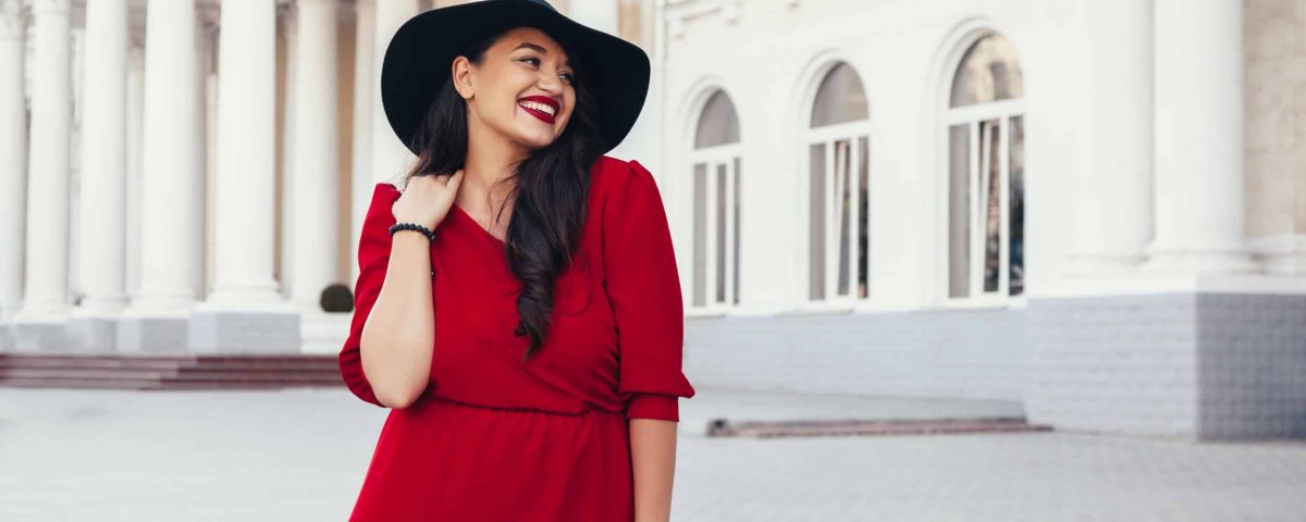 smiling brunette woman with red dress and lipstick outside a mansion
