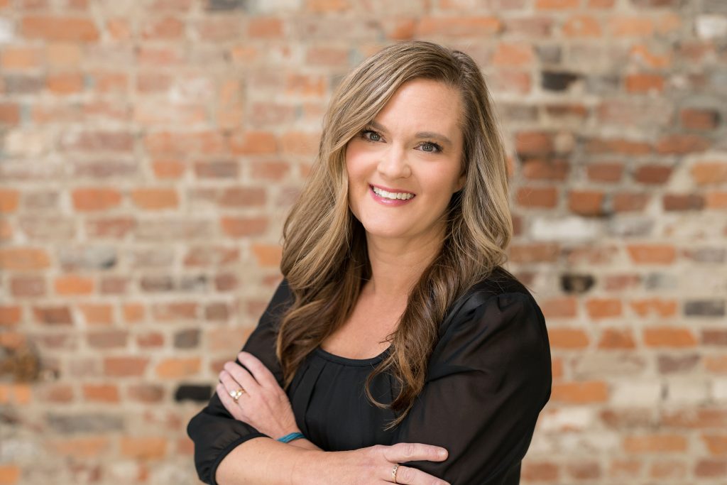Kelly Rice standing with arms folded with brick wall
